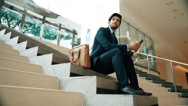 Smart project manager looking at mobile phone while sitting at stairs. Attractive caucasian business man working by using phone. Professional investor using phone to plan business strategy. Exultant.