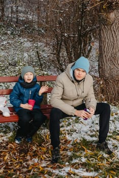 Capture the heartwarming essence of winter as two brothers, aged 8 and 17, share a special moment on a snow-covered bench in a serene rural park. Sipping hot tea from a thermos, they immerse themselves in the beauty of the snowy landscape, creating cherished memories of familial warmth amidst the tranquil winter scenery. This captivating image encapsulates the joy of family bonding, winter relaxation, and the simple pleasures of a cozy picnic in the midst of nature's snowy embrace.