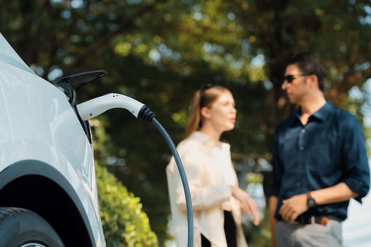 Young couple recharge electric car's battery from charging station in outdoor green city park in springtime. Rechargeable EV car for sustainable environmental friendly urban travel lifestyle.Expedient