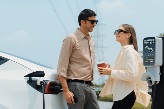 Young couple with coffee recharge EV car battery at charging station connected to electrical industrial power grid tower. Couple with shopping and travel using eco electric car lifestyle. Expedient