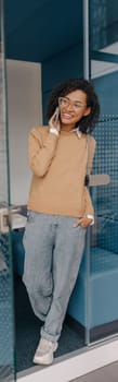 Smiling businesswoman is talking phone with client while standing on modern office background