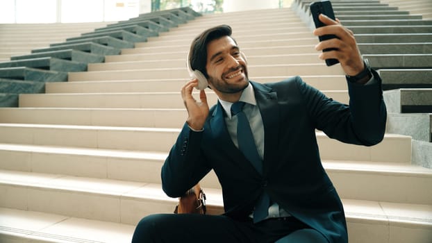 Happy smart business man taking selfie while smiling at smart phone. Closeup image of professional executive manager sitting at stairs while wearing suit and headphone. Creative business. Exultant.