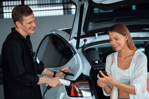 Young couple travel with EV electric car to shopping center parking lot charging in downtown city showing urban sustainability lifestyle by green clean rechargeable energy of electric vehicle innards