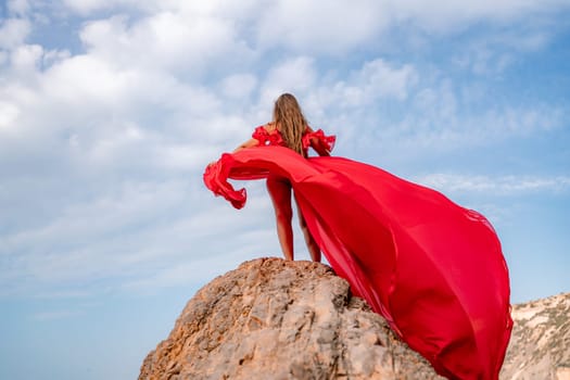 woman sky red dress. Woman with long hair on a sunny seashore in a red flowing dress, back view, silk fabric waving in the wind. Against the backdrop of the blue sky and mountains on the seashore