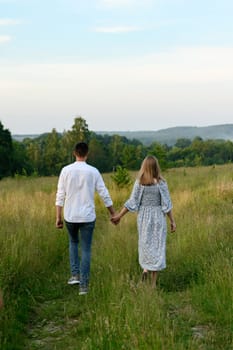 A boy and a girl are holding hands and walking in an open field with the sky and forest on the horizon.