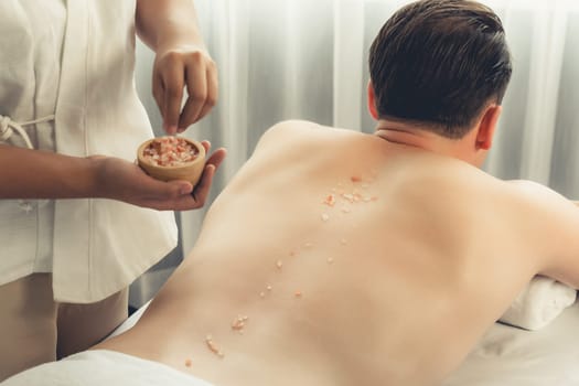 Blissful man customer having exfoliation treatment in luxury spa salon with warmth candle light ambient. Salt scrub beauty treatment in health spa body scrub. Quiescent
