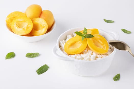 Fresh cottage cheese in white bowl with mint leaves and apricot pieces on white background.