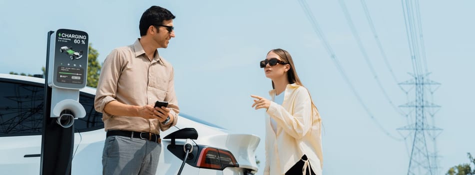 Couple pay for electricity with smartphone while recharge EV car battery at charging station connected to electrical power grid tower as industry for eco friendly car utilization. Panorama Expedient