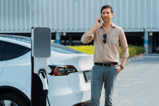 Young man recharge EV electric vehicle at green city commercial mall parking lot while talking on phone. Sustainable urban lifestyle for eco friendly EV car with battery charging station. Expedient