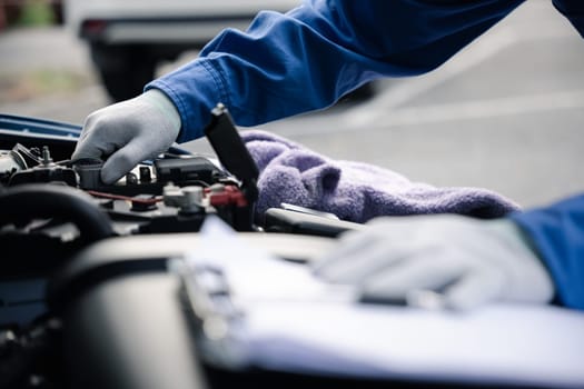 Close-up of an Asian mechanic's hand, in blue workwear, writing down repair notes on a paper while working on a car in an auto repair shop.