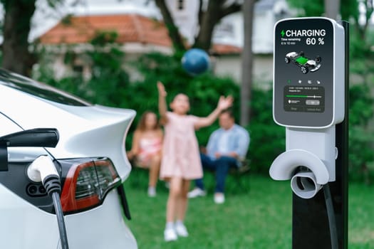 Focus electric vehicle recharge from home charging station on blur background of happy and playful family playing together. EV car using alternative and sustainable energy for better future.Synchronos