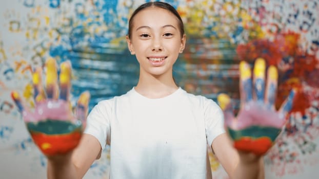 Portrait of happy smart student looking at camera while showing hand to camera with colorful color. Smiling caucasian teenager wearing white shirt while standing at colorful stained wall. Edification.