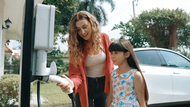 Happy little young girl learn about eco-friendly and energy sustainability as she help her mother recharge electric vehicle from home EV charging station. EV car and modern family concept. Synchronos