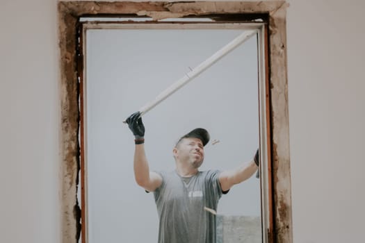 A young caucasian man in a uniform and gray textile gloves with real emotions on his face dismantles a board from a doorway with rubbish pouring onto the floor, close-up view from below with selective focus. Construction work concept.