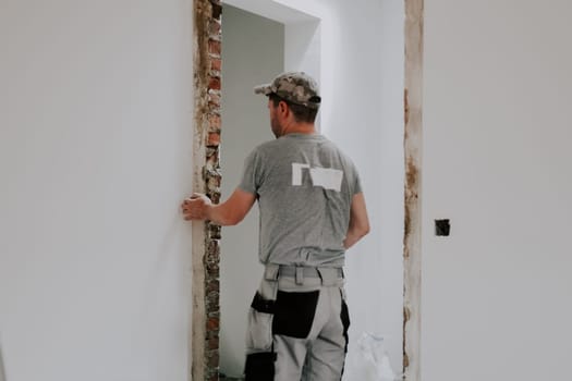 A young Caucasian man in a uniform and gray textile gloves standing with his back brushing off the remnants of debris from a dismantled doorway, close-up side view with selective focus. Construction work concept.