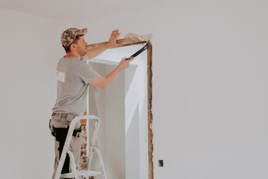 One young Caucasian man in a uniform and cap clears a doorway from old putty using a crowbar while standing on a stepladder in a room with white walls, close-up side view with selective focus. Construction work concept.