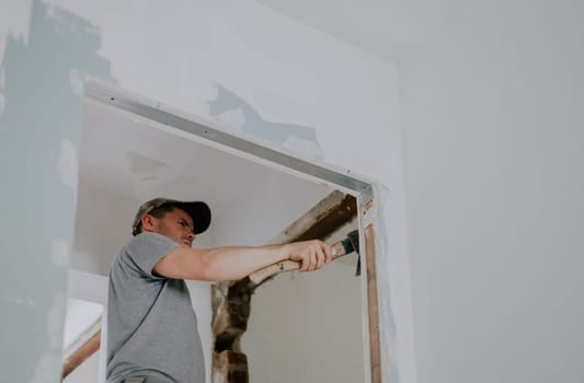 One young Caucasian man in a gray T-shirt and cap clears old putty from the beams of a doorway with one hand using an ax, close-up view from below with selective focus. Construction work concept.