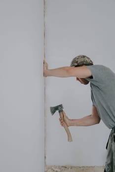 One young Caucasian man in a gray T-shirt and cap is cleaning old putty from the side of a doorway with an ax while standing at an angle, close-up side view with selective focus. Construction work concept.