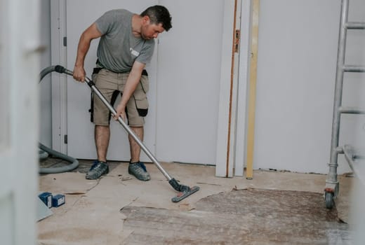 Young caucasian brunette man in work clothes on the left is vacuuming the floor with a construction vacuum cleaner in a room in an old house being renovated, side view close-up with selective focus.Construction work concept.