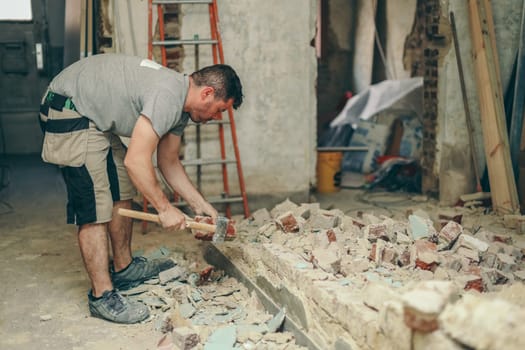 A young caucasian brunette man with a beard in work clothes with a sledgehammer in his hands throws aside a dirty brick from a collapsed wall, close-up side view.Construction work concept.