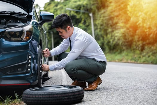 Asian businessman with lug wrench changing car wheel on the highway. Professional car mechanic providing road assistance. Vehicle tyre replacement concept.