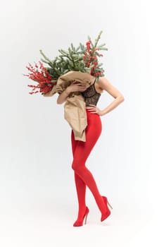 A long girl in red tights and high-heeled shoes holds a huge bouquet of fir branches and red berries wrapped in cardboard paper, she closes with a bouquet, photo studio with white background. High quality 4k footage