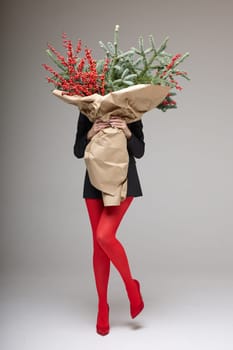 A long girl in red tights and high-heeled shoes holds a huge bouquet of fir branches and red berries wrapped in cardboard paper, she closes with a bouquet, photo studio with white background. High quality 4k footage