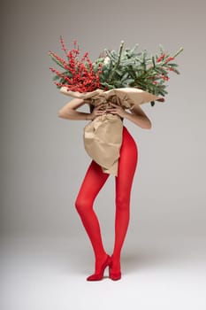 A long girl in red tights and high-heeled shoes holds a huge bouquet of fir branches and red berries wrapped in cardboard paper, she closes with a bouquet, photo studio with white background. High quality 4k footage