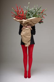 A long girl in red tights and high-heeled shoes holds a huge bouquet of fir branches and red berries wrapped in cardboard paper, she closes with a bouquet, photo studio with white background. High quality 4k footage