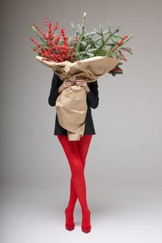 A long girl in red tights and high-heeled shoes holds a huge bouquet of fir branches and red berries wrapped in cardboard paper, she closes with a bouquet, photo studio with white background. High quality 4k footage