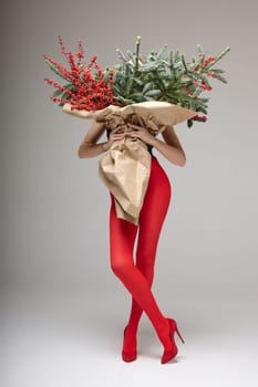 A long girl in red tights and high-heeled shoes holds a huge bouquet of fir branches and red berries wrapped in cardboard paper, she closes with a bouquet, photo studio with white background. High quality 4k footage