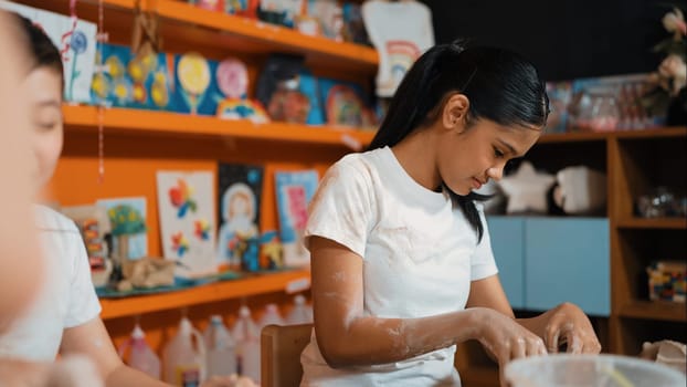 Caucasian highschool girl modeling cup of clay while diverse children laughing in art class at workshop. Multicultural student playing or making clay art in creative activity. Education. Edification.