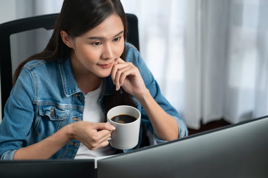 Working young beautiful asian creative manager with glasses focusing on pc while drinking hot coffee, creating with website design on social media with blue jeans shirt at home office. Stratagem.