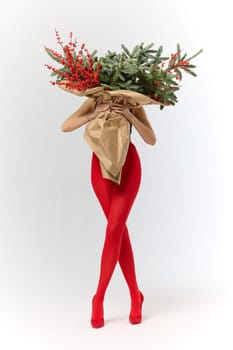 A long girl in red tights and high-heeled shoes holds a huge bouquet of fir branches and red berries wrapped in cardboard paper, she closes with a bouquet, photo studio with white background. High quality 4k footage