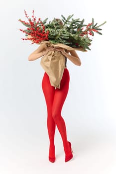 A long girl in red tights and high-heeled shoes holds a huge bouquet of fir branches and red berries wrapped in cardboard paper, she closes with a bouquet, photo studio with white background. High quality 4k footage