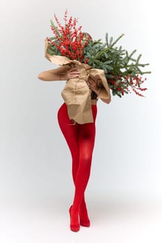 A long girl in red tights and high-heeled shoes holds a huge bouquet of fir branches and red berries wrapped in cardboard paper, she closes with a bouquet, photo studio with white background. High quality 4k footage