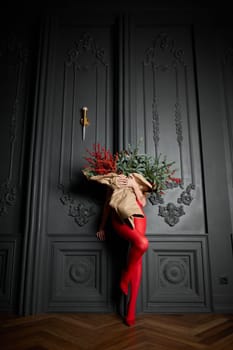 A long girl in red tights and high-heeled shoes holds a huge bouquet of fir branches and red berries wrapped in cardboard paper, she closes with a bouquet, photo studio with grey wall background. High quality 4k footage