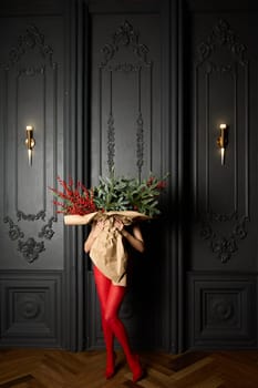 A long girl in red tights and high-heeled shoes holds a huge bouquet of fir branches and red berries wrapped in cardboard paper, she closes with a bouquet, photo studio with grey wall background. High quality 4k footage
