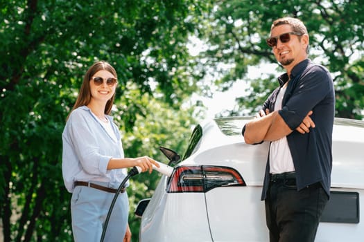 Lovely young couple wearing sun glasses recharging battery for electric car during road trip travel EV car in natural forest or national park. Eco friendly travel during vacation and holiday. Exalt