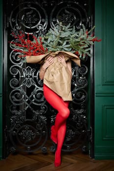 A long girl in red tights and high-heeled shoes holds a huge bouquet of fir branches and red berries wrapped in cardboard paper, she closes with a bouquet, photo studio with black grill background. High quality 4k footage