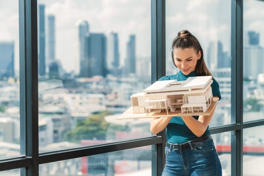 Beautiful young architect engineer holds architectural model while inspect house model. Professional interior designer checking house construction while standing near window with city view. Tracery.