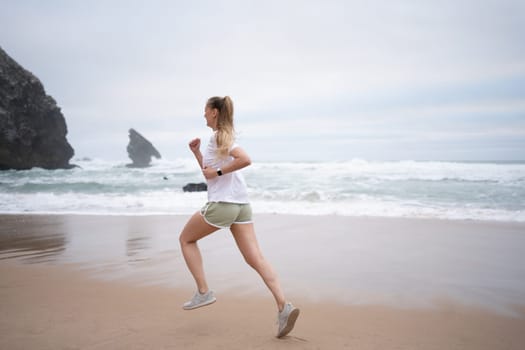 Full length of athletic woman running along seashre. Young sportswoman in white top and sporty shorts jogging on sandy beach. Fit lady represents healthy lifestyle.