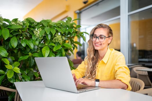 Smiling young female freelancer using laptop at cafe table. Woman in yellow jacket with blond hair sitting near green plant on cafe terrace. Remote working concept.