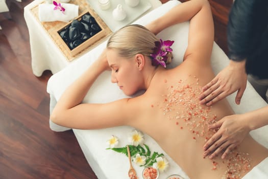 Panorama top view woman customer having exfoliation treatment in luxury spa salon with warmth candle light ambient. Salt scrub beauty treatment in health spa body scrub. Quiescent