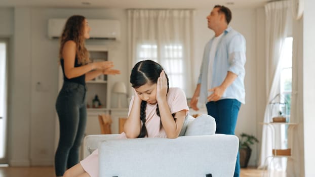 Annoyed and unhappy young girl sitting on sofa trapped in middle of tension by her parent argument in living room. Unhealthy domestic lifestyle and traumatic childhood develop to depression Synchronos