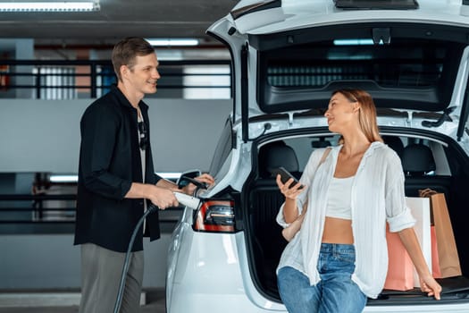 Young couple travel with EV electric car to shopping center parking lot charging in downtown city showing urban sustainability lifestyle by green clean rechargeable energy of electric vehicle innards