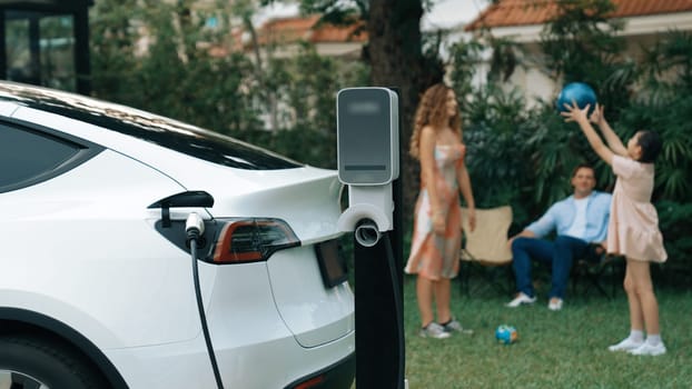 Electric vehicle recharge from home charging station on background of happy and playful family playing together. EV car using alternative and sustainable energy for better future Synchronos