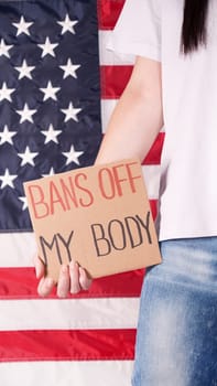 Woman holding a sign Bans Off My Body American flag on background. Protest against anti abortion law. Women's strike. Womens rights freedom