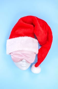 Pink sleeping eye mask on mannequin face with red christmas hat on blue background, sleeping disorder. Holidays, Head accessory. Last minute shopping sickness, Plastic face