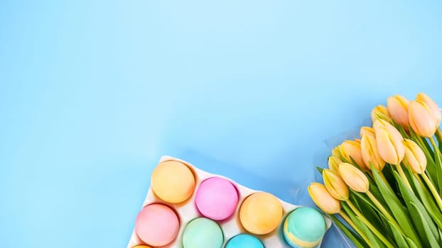 Colorful Easter eggs pack and yellow tulips on blue background with space for text. Spring flat lay. Pastel painted eggs in box